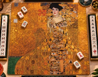Op de afbeelding: Een mahjongset met een speelbord bedrukt met Gustav Klimt's Portret van Adele Bloch-Bauer I. De set bevat vier tegelhouders, witte tegels met rode, groene en zwarte symbolen, een cocktailglas en decoratieve bloemstukken.