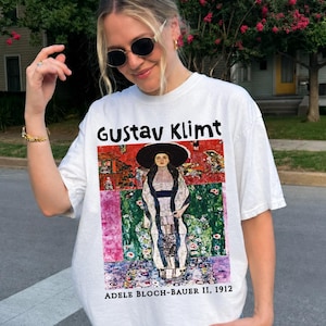 May include: A white t-shirt featuring a graphic print of the Gustav Klimt painting Adele Bloch-Bauer II, 1912. The artwork depicts a figure in a wide-brimmed black hat and patterned clothing against a colorful background. The text Gustav Klimt and Adele Bloch-Bauer II, 1912 is printed in black.