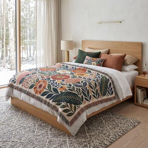 Puede incluir: Una manta tejida con fondo crema y un patr&oacute;n repetitivo de flores grandes en tonos naranja, rojo y amarillo, hojas verde oscuro y bayas peque&ntilde;as. La manta tiene un borde con flecos y est&aacute; extendida sobre una cama con ropa blanca.