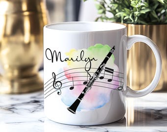 Op de afbeelding: Witte keramische mok met een zwarte klarinet, muzieknoten en de naam "Marilyn" in script. Het ontwerp bevat een kleurrijke aquarel achtergrond met gele, groene, blauwe en roze tinten. De mok heeft een gebogen handvat.