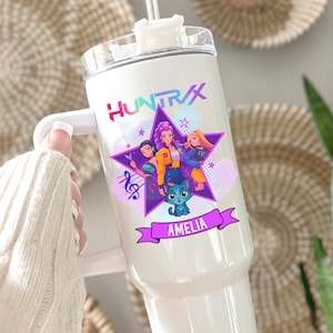 May include: White insulated tumbler with a handle, featuring a star design with three cartoon figures and a blue cat. The tumbler has the text "HUNTRAX" and the name "AMELIA" on a banner.