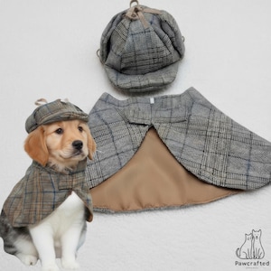 Grey plaid detective pet costume laid flat, the deerstalker hat above the hound cape which is opened to show its camel satin lining, beside a golden and white puppy wearing the same set.