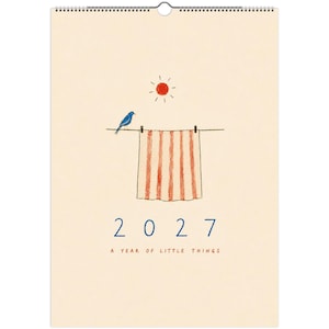 May include: A wall calendar with a cream background featuring a blue bird perched on a clothesline next to a red and white striped towel hanging from two clips. Above is a red sun. The text reads 2027 A YEAR OF LITTLE THINGS in blue and red.