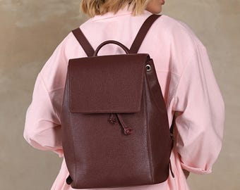 May include: A burgundy leather backpack with a flap closure and drawstring detail. The backpack has adjustable straps and a top handle. The bag is shown from the back, with the straps over the shoulders.