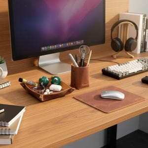 Può includere: Un'organizzazione da scrivania ben organizzata con un monitor del computer, una tastiera e delle cuffie. Un vassoio in pelle marrone contiene piccoli oggetti e un portapenne abbinato è nelle vicinanze. Sono visibili anche un mouse e un tappetino.