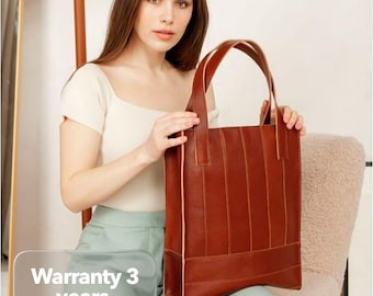 May include: A brown leather tote bag with vertical stitching and a rectangular shape. The bag has two handles and is held by a person. The text "Warranty 3 years" is visible in the image.