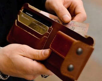 May include: A brown leather wallet with multiple card slots and a bill compartment. The wallet is open, revealing credit cards and paper currency. The wallet has a snap closure and is held by a person.