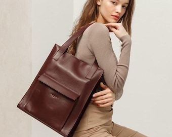 May include: A burgundy leather tote bag with a rectangular shape and a front pocket. The bag has two handles and is being held by a person. The bag is a deep red color and appears to be made of high-quality leather.