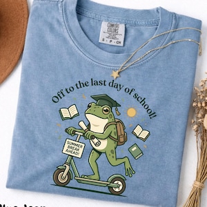 May include: Blue jean colored t-shirt with a graphic of a frog riding a scooter, wearing a graduation cap and backpack. The text reads "Off to the last day of school!" and "Summer Break Ahead!"