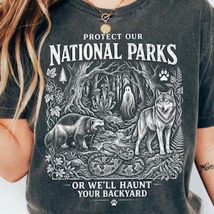 Peut inclure: T-shirt de couleur poivre avec un motif "Protect Our National Parks" au-dessus d'une scène forestière détaillée avec un loup, un blaireau, un serpent et un fantôme. Le texte "Or We'll Haunt Your Backyard" est en dessous.