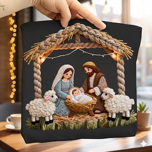 May include: Black square pillow featuring an embroidered Nativity scene. The scene includes Mary, Joseph, baby Jesus in a manger, and two sheep. The stable is made of woven straw and rope, with string lights.
