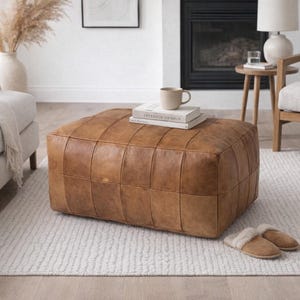 May include: A large, square, brown leather ottoman with a grid pattern sits on a white rug. A stack of books and a mug rest on top. The ottoman is a warm, neutral color, and the room has a cozy, inviting atmosphere.