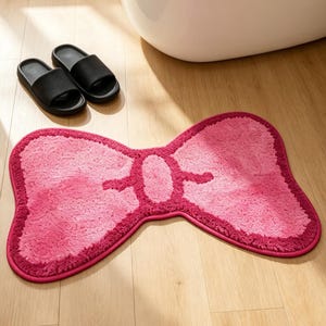 Può includere: Un tappetino da bagno rosa a forma di fiocco con un bordo rosa più scuro su un pavimento in legno chiaro. Due sandali neri sono vicino al tappetino. Sullo sfondo c'è una vasca da bagno bianca.