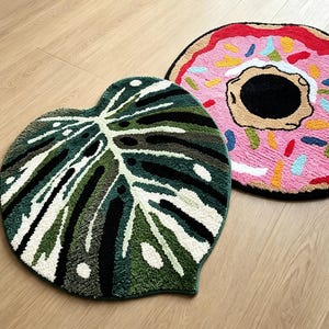 Op de afbeelding: Twee getufte vloerkleden met verschillende ontwerpen. Het ene kleed heeft de vorm van een monstera-blad in tinten donkergroen, lichtgroen en wit. Het andere kleed is rond en lijkt op een donut met roze glazuur, kleurrijke hagelslag en zwart.
