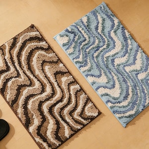 Könnte beinhalten: Zwei rechteckige Badematten mit einem wellenförmigen Muster. Eine Matte ist braun und beige, die andere blau und weiß. Die Matten bestehen aus einem weichen, strukturierten Material und liegen auf einem hellbraunen Holzboden.
