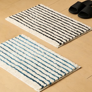 May include: Two rectangular bath mats with striped patterns. One mat has black stripes on a white background, and the other has blue stripes on a white background. Black sandals are placed on the wooden floor near the mats.