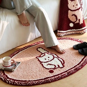 Puede incluir: Una alfombra de forma ovalada con una superficie texturizada de color rosa claro que presenta un gráfico de un gato blanco con contornos rojo oscuro. La alfombra tiene un borde rojo oscuro con detalles en blanco.