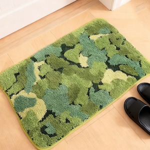 May include: A rectangular bath mat with a camouflage pattern in shades of green, with a light green border. Two black rubber sandals are next to the mat on a wooden floor.