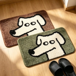 Puede incluir: Dos felpudos rectangulares con temática de perros. Uno es marrón, el otro verde. Cada alfombra presenta una cara de perro blanca con contornos negros. Sandalias negras colocadas cerca de las alfombras sobre un suelo de madera.