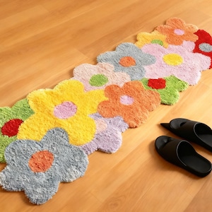 Peut inclure: Un long tapis touffeté rectangulaire présentant un motif répétitif de fleurs interconnectées dans diverses couleurs, notamment jaune, orange, rouge, bleu clair, rose, vert et lavande. Une paire de sandales noires est posée à côté du tapis.