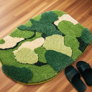 Puede incluir: Una alfombra de baño verde con estampado de camuflaje en varios tonos de verde y beige. La alfombra tiene un aspecto suave y texturizado y está colocada sobre un suelo de madera. Dos sandalias negras están al lado de la alfombra.
