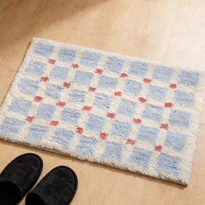 May include: A rectangular bath mat featuring a pattern of light blue squares and small red dots on an off-white background. Two black slippers are at the bottom of the image. The mat is made of a soft, absorbent material.