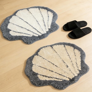 May include: Two seashell-shaped bath mats with a white and beige design, outlined in gray. A pair of black open-toe slippers are also visible. The mats are made of a soft, textured material.
