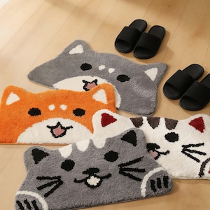May include: Several cat-shaped rugs in shades of orange, grey, and white, with black facial features. Two pairs of black slide sandals are also visible. The rugs are arranged on a light wooden floor.