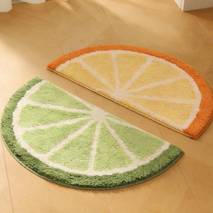 以下が含まれることがあります： 2つの半円形のバスマット。1つはオレンジ色の皮が付いた黄色で、レモンスライスに似ています。もう1つは白い内側が緑色で、ライムスライスに似ています。どちらのマットも明るい茶色の木の床にあります。