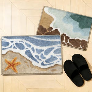May include: Two rectangular bath mats with beach-themed designs. One features a blue ocean wave with a starfish, the other a brown and green coastal scene. Black slippers are placed nearby. The mats have a beige border.