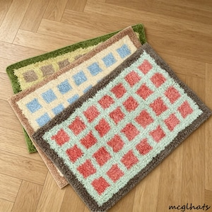 May include: Three rectangular tufted rugs with a grid pattern. The top rug is green with tan squares, the middle is cream with light blue squares, and the bottom is mint green with pink squares. Each rug features a solid colored border.
