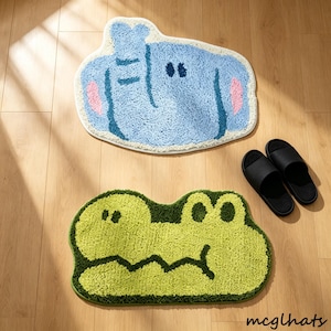 May include: Two cartoon bath mats, one blue elephant and one green alligator, are on a wooden floor. The elephant mat has pink ears and the alligator mat has dark green details. A pair of black sandals is next to the mats.