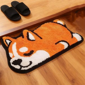 Puede incluir: Una alfombra de baño novedosa con forma de perro Shiba Inu, con colores naranja, blanco y negro. La alfombra está sobre un suelo de madera, junto a un par de zapatillas negras. La alfombra tiene una superficie suave y texturizada.