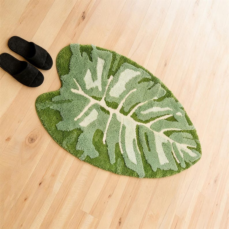 May include: A green leaf-shaped bath mat with a lighter green and white leaf pattern. Two black slippers are placed to the left of the mat on a light wood floor. The mat is approximately 91 cm long.