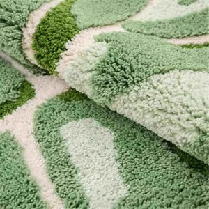 May include: Close-up of a soft, textured bath mat with a leaf-like pattern. The mat features shades of green and white, creating a natural, organic design. The plush material suggests comfort and absorbency, ideal for a bathroom setting.