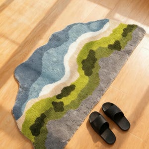 Op de afbeelding: Een abstracte badmat met een bergketenontwerp in blauw, groen en grijs. Twee zwarte sandalen liggen naast de mat op een houten vloer. Het ontwerp van de mat lijkt op een landschap.