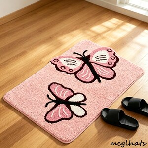 May include: A rectangular pink bath mat with a textured, plush surface featuring two butterfly designs. Each butterfly has dark brown bodies and outlines with light pink and white wing patterns. The text mcglhats is printed in the bottom corner.