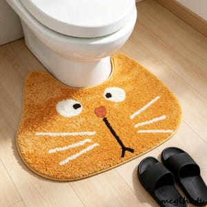 Puede incluir: Una alfombra de baño de felpa naranja con forma de gato y un recorte para la base del inodoro. La alfombra tiene ojos bordados blancos con pupilas negras, una pequeña nariz rosa y bigotes blancos cosidos a ambos lados de una boca oscura.