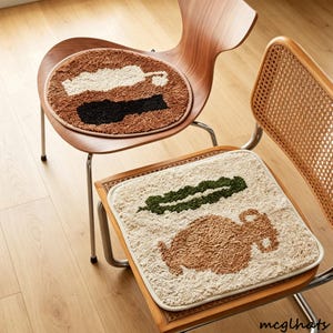 Könnte beinhalten: Zwei getuftete Sitzkissen auf Holzstühlen. Das runde Kissen zeigt braune, weiße und schwarze abstrakte Formen. Das quadratische Kissen ist cremefarben mit einem dunkelgrünen blattartigen Design und einer braunen, texturierten, amphorenförmigen Grafik.