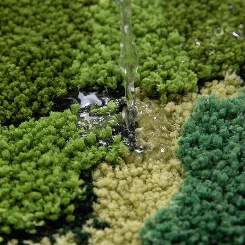 May include: Close-up of a green, textured bath mat with water pouring onto it. The mat has varying shades of green, creating a textured, grassy appearance. Water droplets are visible on the surface, highlighting the mat's absorbent qualities.
