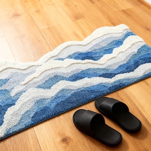 May include: A blue and white mountain-shaped bath mat with a textured design. The mat is placed on a wooden floor next to a pair of black sandals.