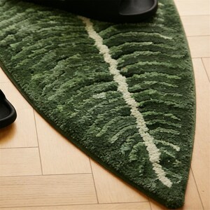 May include: A green leaf-shaped rug with a white vein design. The rug is made of a soft, textured material and is placed on a light-colored wooden floor. Black sandals are partially visible on the rug.