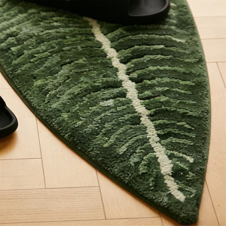 May include: A green leaf-shaped rug with a white vein design. The rug is made of a soft, textured material and is placed on a light-colored wooden floor. Black sandals are partially visible on the rug.