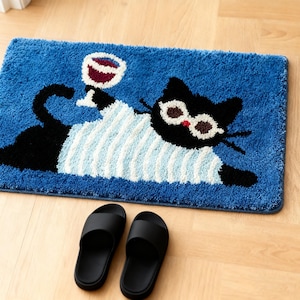 Puede incluir: Una alfombra de baño azul con un diseño de gato negro con gafas y una camisa a rayas, sosteniendo una copa de vino. Dos chanclas negras están frente a la alfombra. El diseño del gato es en blanco, negro y rojo.