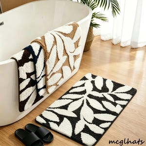 May include: A bathroom set featuring a black and white leaf-patterned bath mat and three matching towels in brown, white, and navy blue. The towels are draped over the edge of a white bathtub. Black sandals are on the wooden floor.