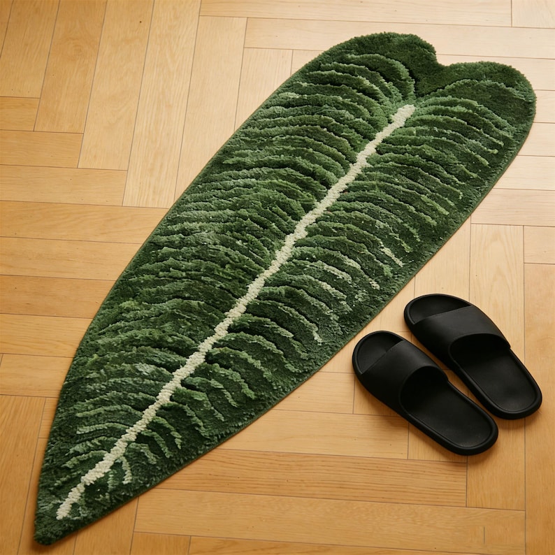 May include: A green leaf-shaped rug with a white central vein, placed on a wooden floor. Two black sandals are next to the rug. The rug's design mimics a large tropical leaf.