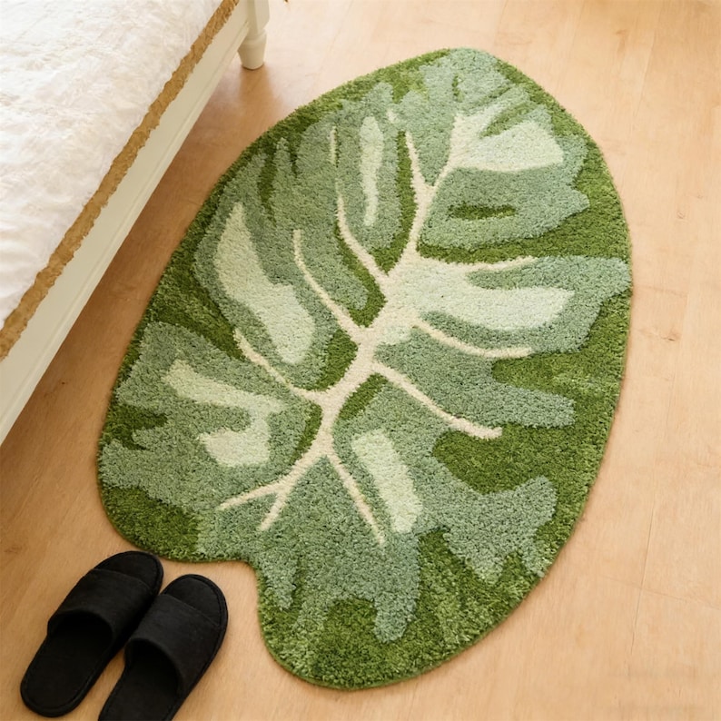 May include: A green leaf-shaped rug with varying shades of green and a cream-coloured vein pattern. The rug is placed on a light-coloured wooden floor, with a pair of black slippers nearby. The rug is near a bed.