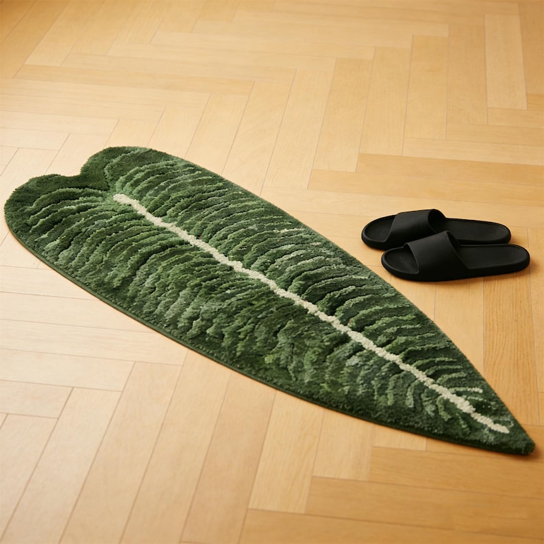 May include: A green leaf-shaped bath mat with a white central vein and textured details. The mat is next to a pair of black slide sandals on a light wood floor. The rug is approximately 60 inches long.