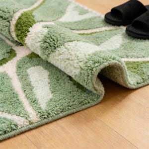 May include: A rolled-up bath mat featuring a green and white leaf pattern. The mat is made of a soft, plush material. Black sandals are visible in the background, resting on a wooden floor.