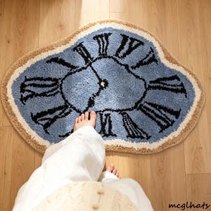 Può includere: Un tappeto tufted dalla forma irregolare con un centro azzurro e numeri romani neri disposti a quadrante d'orologio. Il tappeto presenta un bordo beige spesso e testurizzato con una struttura a pelo morbido e soffice dall'aspetto distorto.